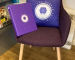 A photo of a purple chair in a library that has a purple cushion and purple ring binder folder on it. Both the cushion and folder have a logo on that says 'The Purple Chair' with a picture of a purple chair on it.