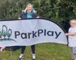 Becky (mum) and Lottie (daughter) stood by a large ParkPlay flag, smiling at camera.
