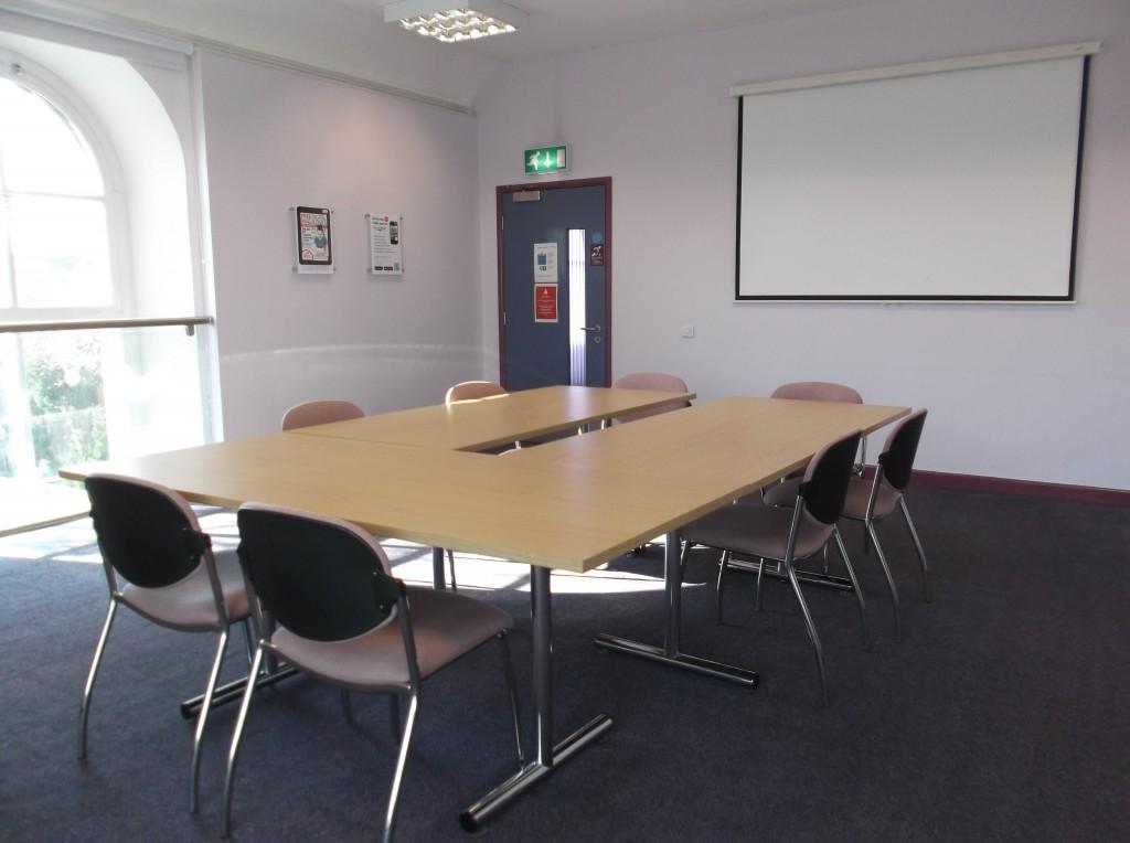 An interior meeting room with grey carpet, pink chairs and a table, and a whiteboard on the wall