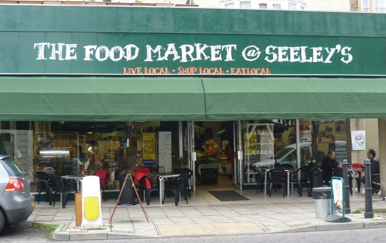 A green shop front with a sign over the window that says The Food Market at Seeleys, with tables out front