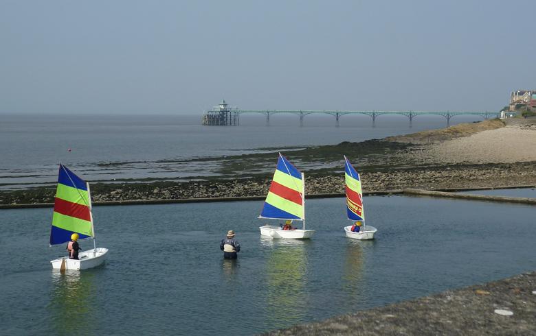 Three small boats with striped sails float on a small lake, with the Clevedon Pier in the distance