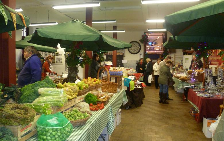 An interior view of a food market, with vegetables set up on a table and green umbrellas over the tables.