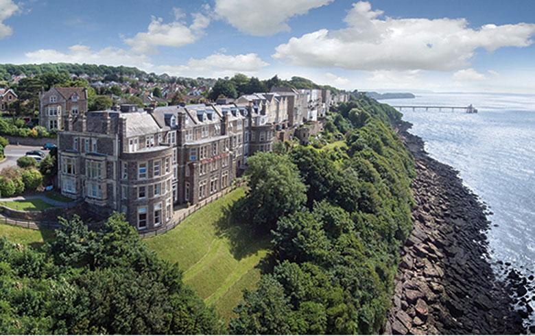 A large Victorian stone hotel sits on a cliff overlooking the sea. 