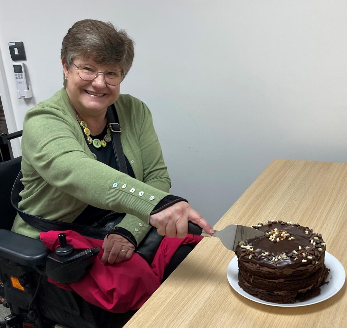A photo of a lady in a wheelchair smiling at the camera while cutting into a large chocolate cake.