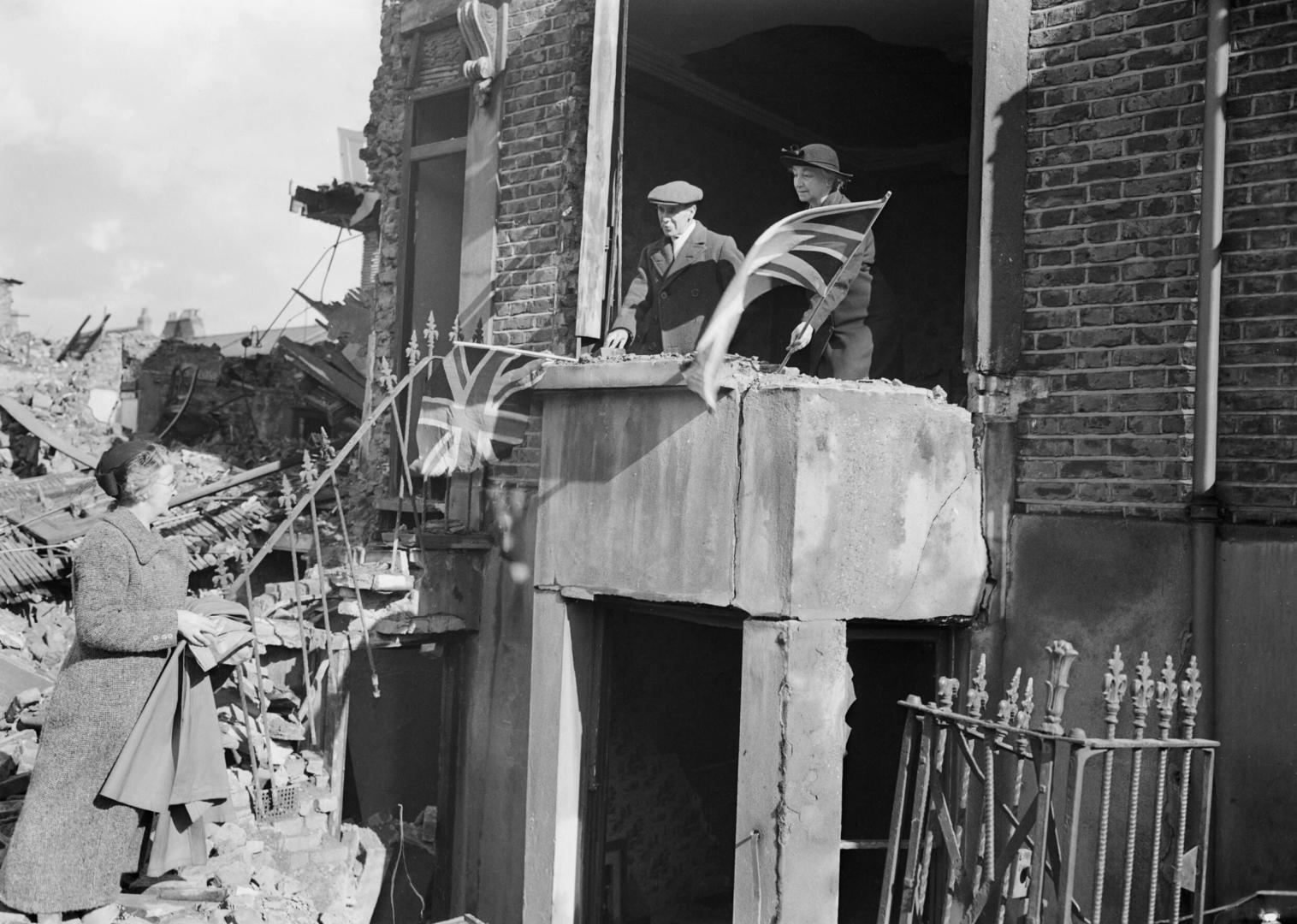 A black and white photo showing bomb damage caused to British houses with people talking and waving the Union Jack flag