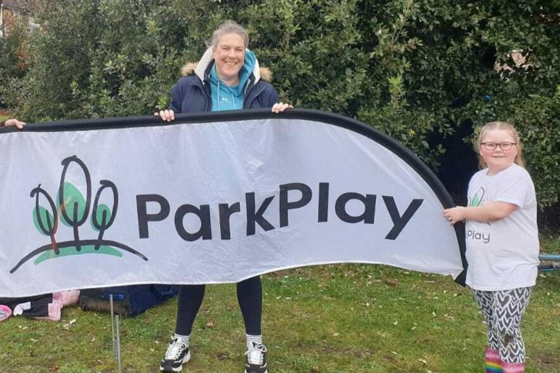 Becky (mum) and Lottie (daughter) stood by a large ParkPlay flag, smiling at camera.