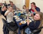 A  group of two adults and four children sat around a table enjoying a meal, they are all smiling and doing a thumbs up to the camera