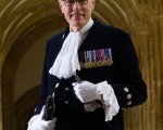 A photo of a white man in glasses with grey hair standing in front of the arches of a church. He is wearing the uniform of the high sheriff with white cravat and gloves, and has medals on his chest. 