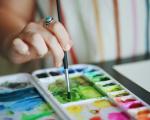 A person dips their paintbrush into green paint on a colourful watercolour palette