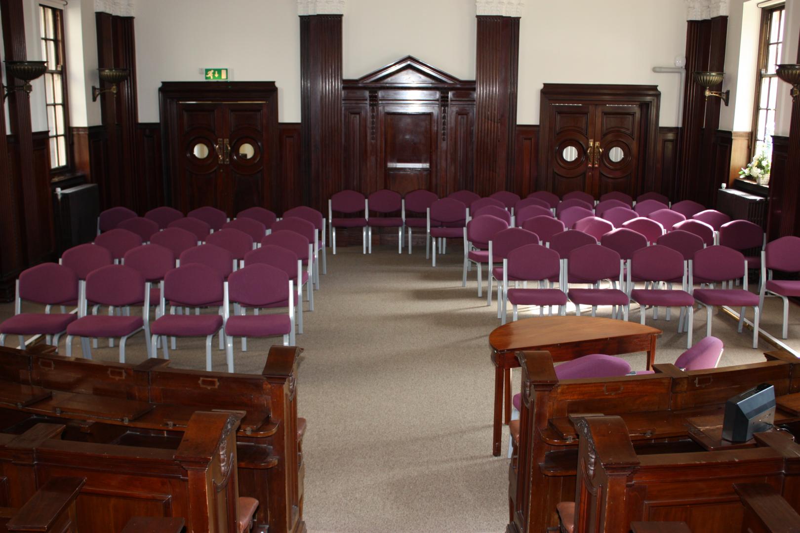 An overview of an oak panelled room, with wooden pews left and right, and pink office chairs lined up on the other side.