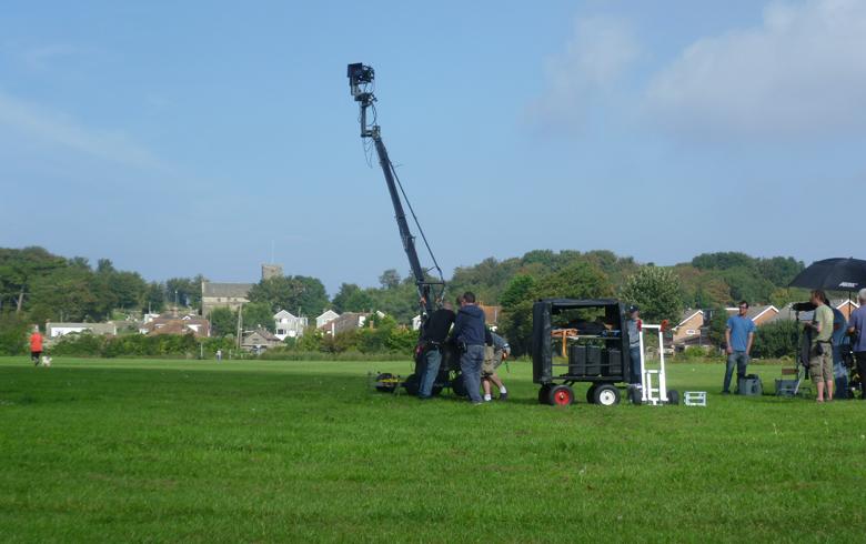 A tall camera crane is surrounded by people in a large open green field