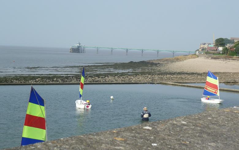 Three small boats with striped sails float on a small lake, with Clevedon Pier in the background