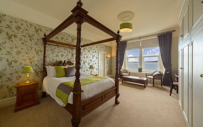 An interior view of a hotel bedroom with a four poster bed, green blankets and floral wallpaper, with a window and view of the sea