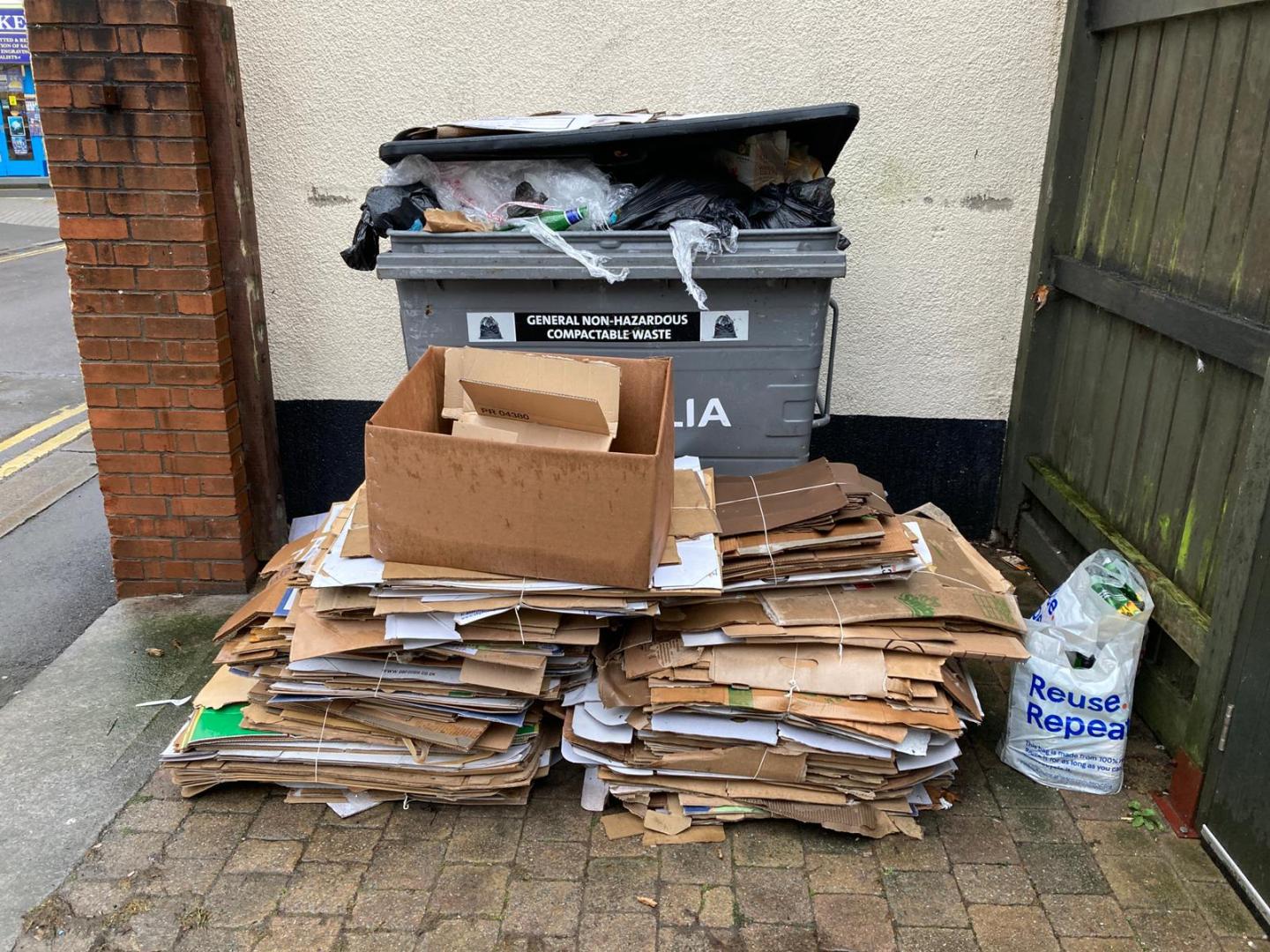 A photo of a large business waste container overflowing with rubbish and a large pile of cardboard in front