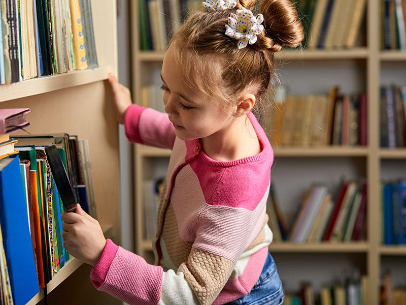 young girl with a magnifying glass inspecting books on a shelf