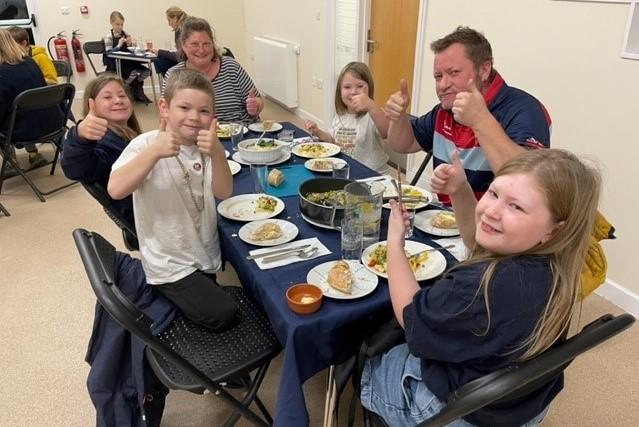 A  group of two adults and four children sat around a table enjoying a meal, they are all smiling and doing a thumbs up to the camera