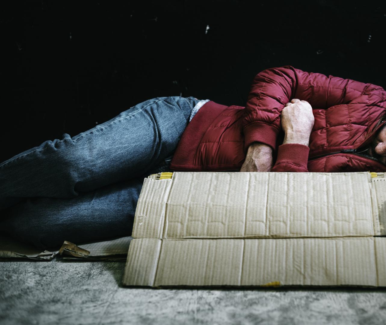 homeless white man wearing blue jeans and a read puffer jacket sleeping out in the cold (rawpixel.com)