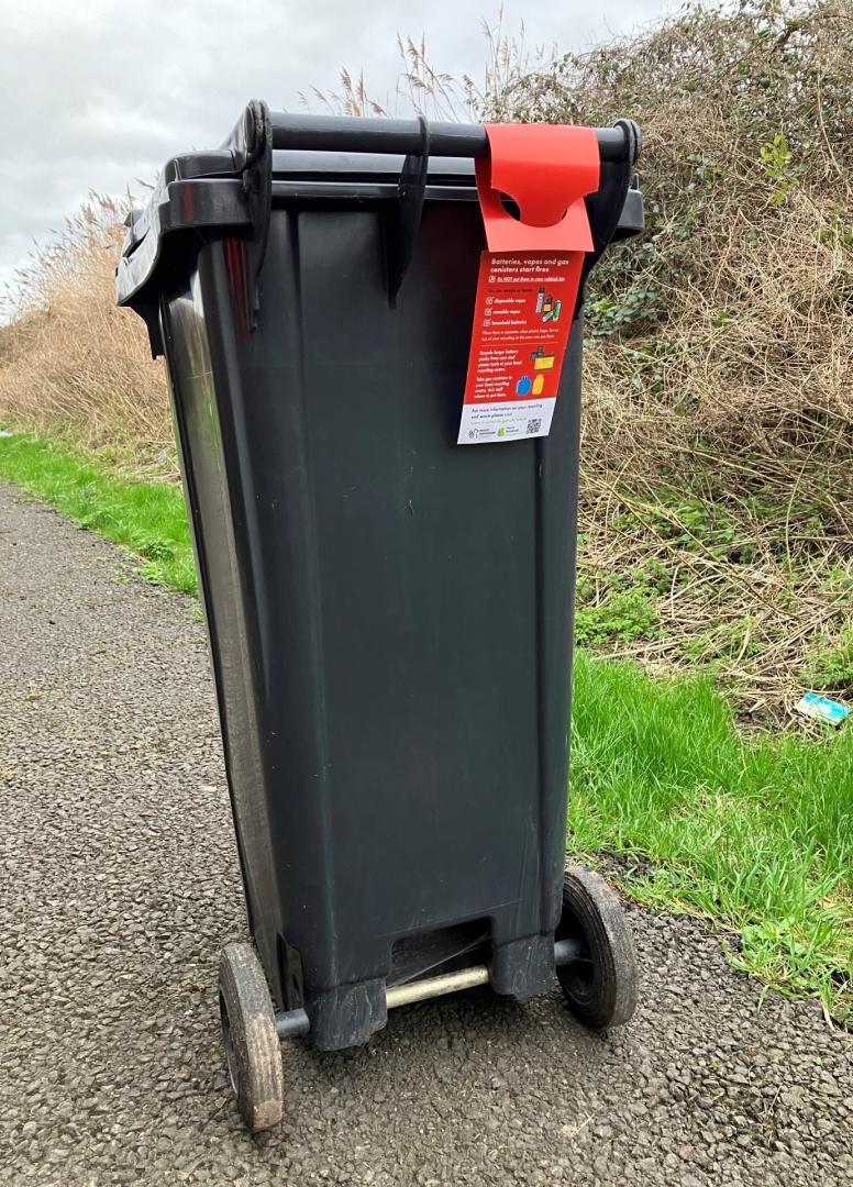 A red hanger explaining how to dispose of batteries safely is attached to the handle of a black general waste bin. There is a hedgerow in the background.