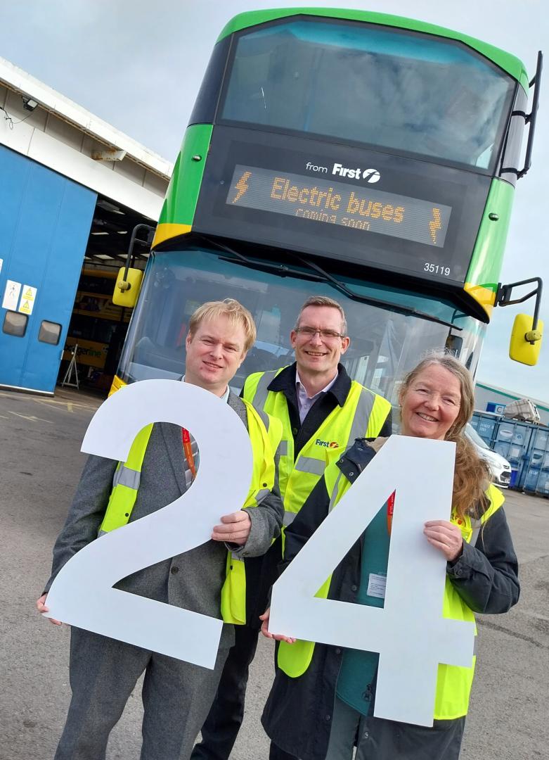 Three people holding giant 24 celebrating electric buses are coming