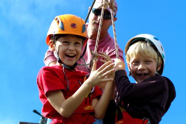 Two children with hardhats on smiling whilst hanging from a harness.
