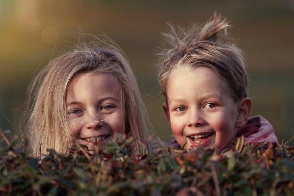 Two young children peeking over a bush and smiling.